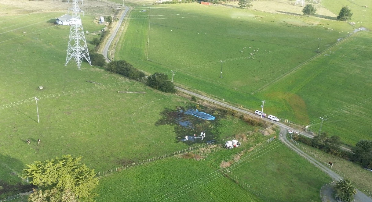 Aerial photographs showing investigation work at the site of the leak. This is the same field from Figure 4 and in the same area which appeared to have had earthworks completed. The pylon, road, and house are all visible as landmarks. There is a large area where the fuel has soaked the ground causing it to turn a black colour. In some areas a neon blue liquid has formed pools, this appears to be areas with a higher concentration of jet fuel. Aerial photographs showing investigation work at the site of the leak. This is the same field from Figure 4 and in the same area which appeared to have had earthworks completed. The pylon, road, and house are all visible as landmarks. There is a large area where the fuel has soaked the ground causing it to turn a black colour. In some areas a neon blue liquid has formed pools, this appears to be areas with a higher concentration of jet fuel.